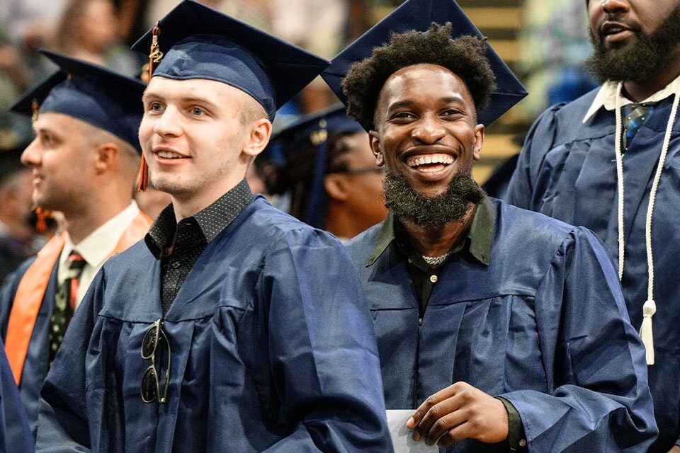 Three male Utica graduates smiling towards the crowed during graduation with blue cap and gowns on.