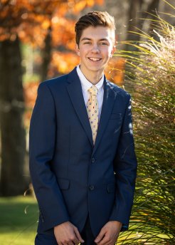 Jack Hrustich III in a blue suit, yellow tie and white shirt, stands outside and smiles.