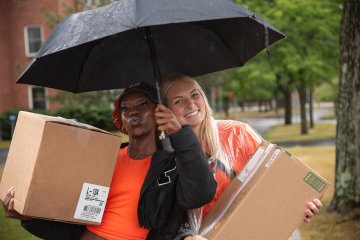 Students under an umbrella hold boxes and smile for the camera.
