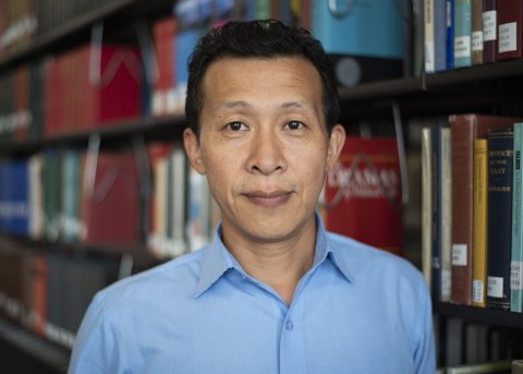 Alanpou Noel Newme in blue button down shirt stands in front of library stacks.