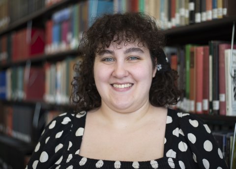 Gabby Hudziak in black and white polka dot shirt stands in front of library stacks.