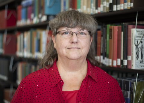 Lisa Rogers in glasses and red shirt stands in front of library stacks.