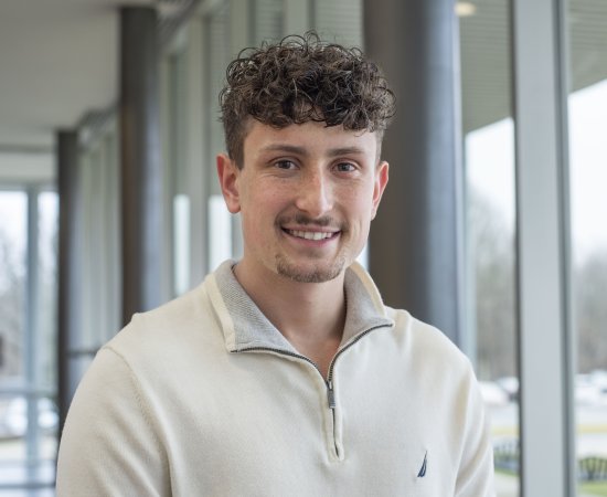 Conner Crossley, in white zip-up pullover, stands by a window in Bull Hall and smiles.