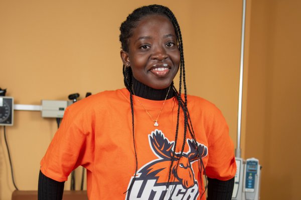 Lois Mensah, in an orange Utica t-shirt, stands against an examination table in the nursing lab.