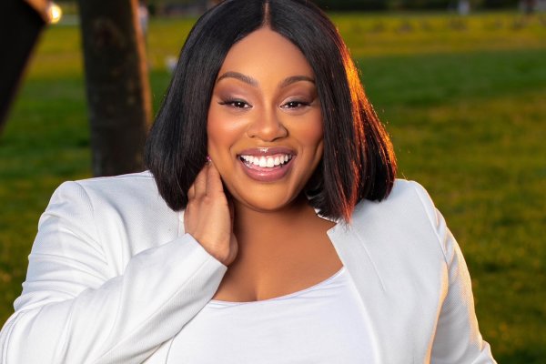 Akira Armstrong, in a white top and white blazer, stands outside on green grass, with her hand held up to her neck and smiles.