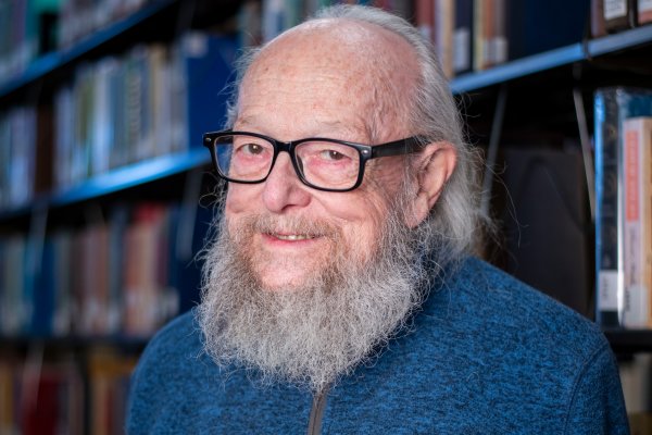 John Cormican, with gray beard and hair down to his shoulders, wearing black glasses and a blue sweater, stands in front of book shelves and smiles.