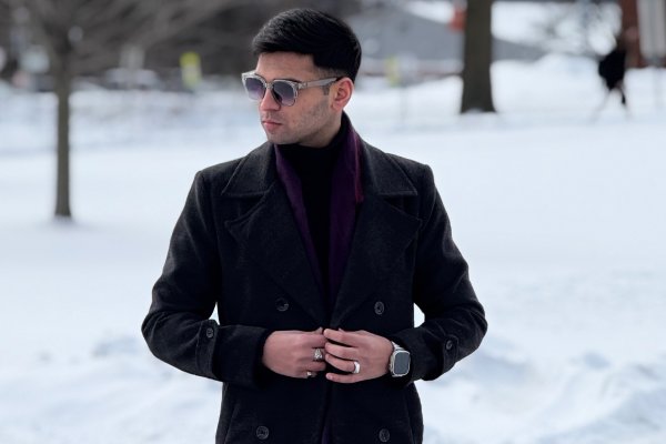 Muhammad Hamza Bilal in sunglasses, buttons up black coat while walking against a snowy backdrop.