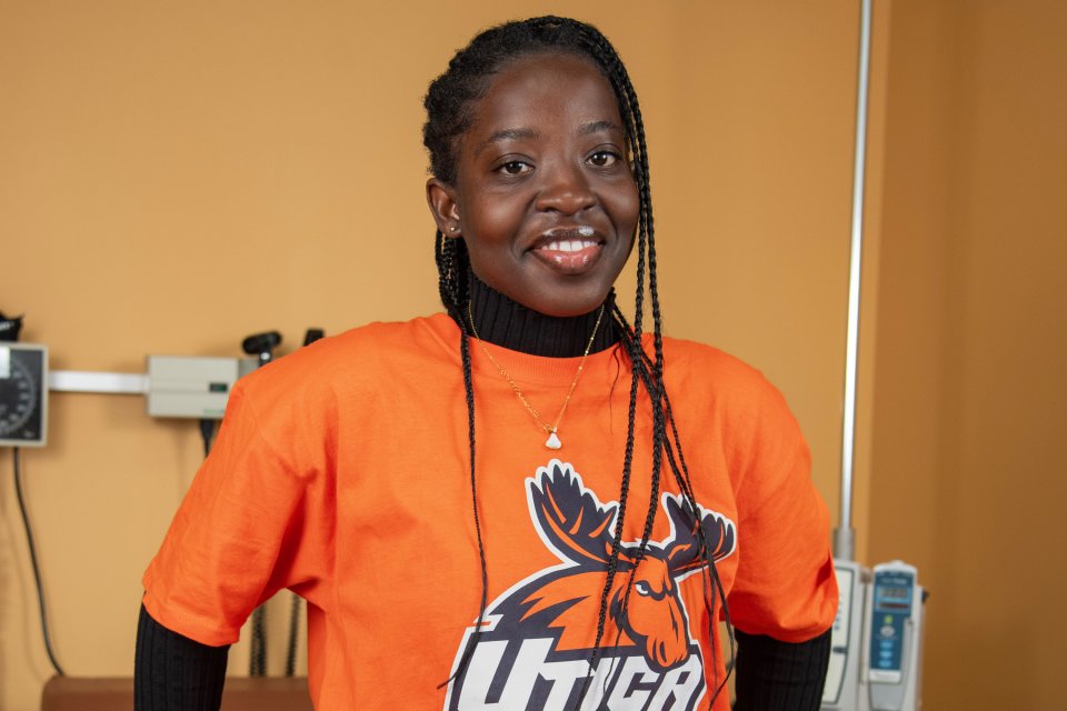 Lois Mensah, in an orange Utica t-shirt, stands against an examination table in the nursing lab.
